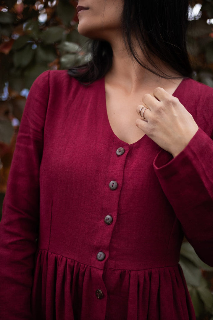 A woman wearing a red dress with a button-down front and long sleeves stands in front of a blurred background of greenery.