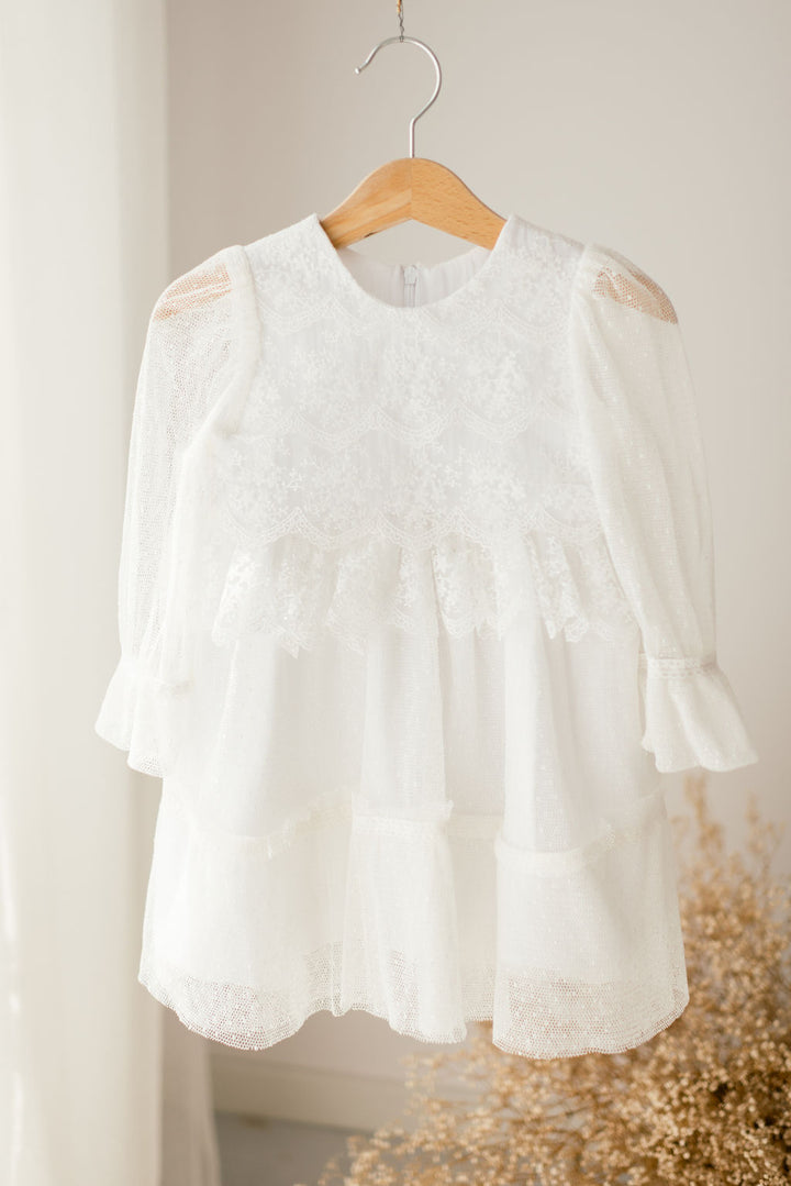 A white lace dress with ruffled sleeves and a ruffled hem hangs on a wooden hanger against a white wall.