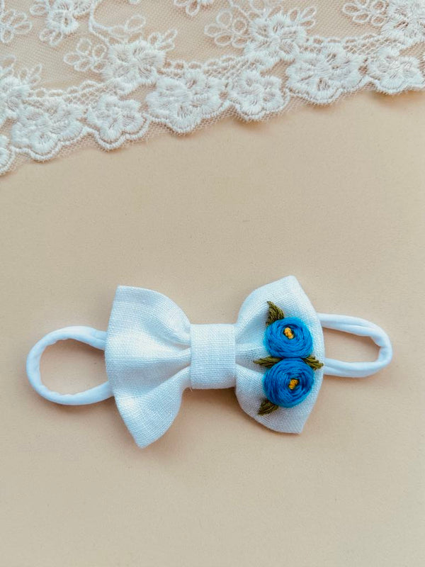 A white bow with blue flowers is placed on a beige background, with a lace doily visible in the top left corner.