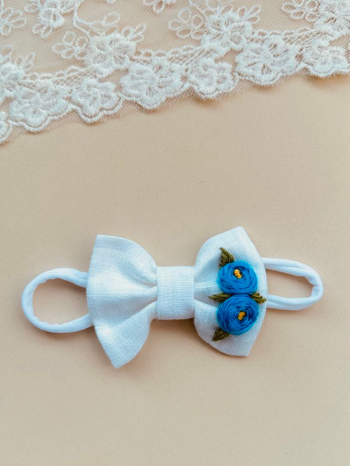 A white bow with blue flowers is placed on a beige background, with a lace doily visible in the top left corner.