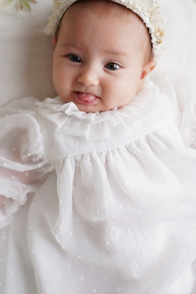 a baby in a white dress laying on a white blanket