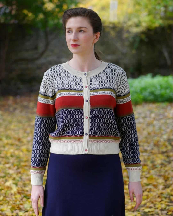 A woman wearing a colorful cardigan and skirt stands in a park surrounded by fallen leaves.