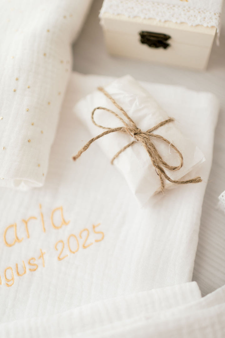 a white cloth with the text "Aria August 2025" embroidered on it, along with a small wrapped item tied with twine and a small white box with a lace-like top.