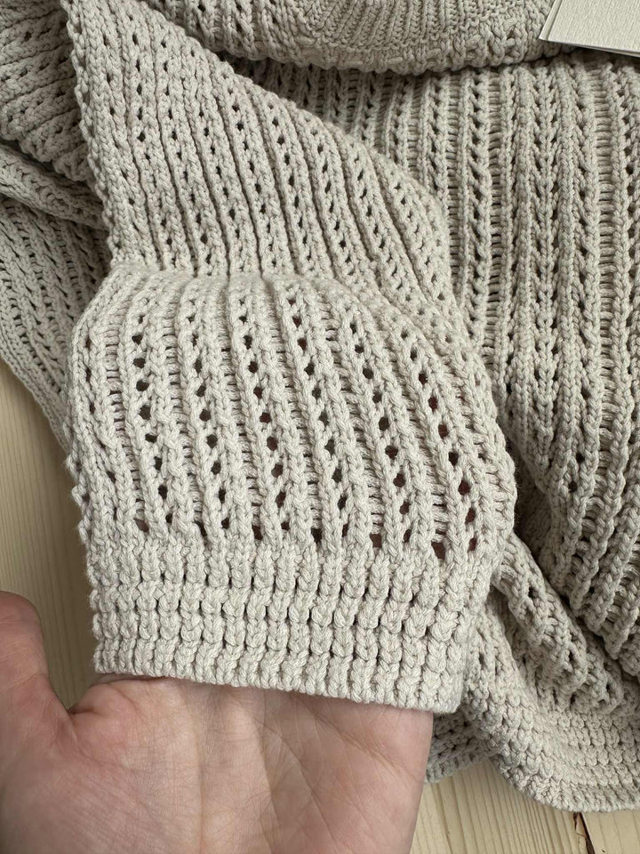 A close-up view of a hand holding a beige knitted or crocheted fabric, with a textured and intricate pattern visible.