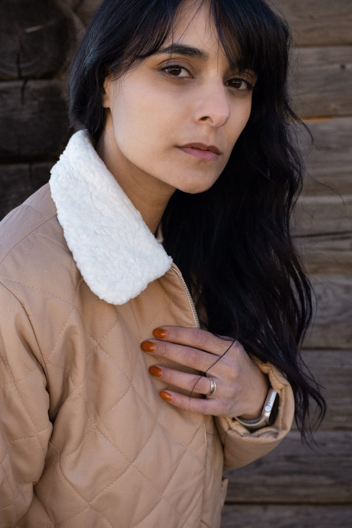 A woman with long dark hair wearing a beige jacket with a white fur collar stands in front of a wooden wall.