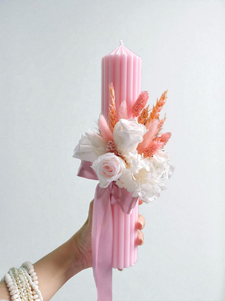 A hand holding a pink candle with a floral arrangement on top.