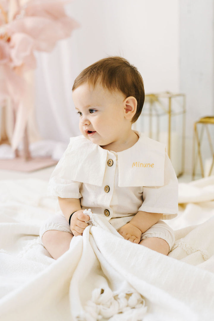 A baby wearing a white outfit with a name tag sits on a white blanket, looking off to the side.