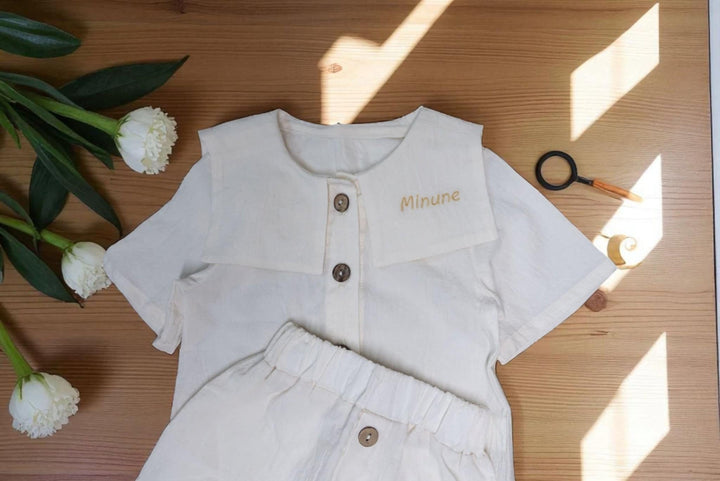 a white outfit with a name tag, placed on a wooden surface alongside some flowers and a magnifying glass.