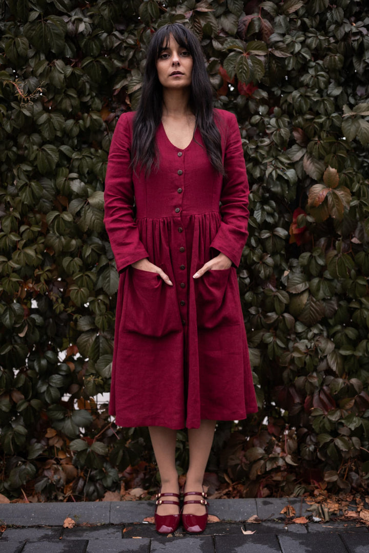 A woman in a red dress stands in front of a green hedge, with her hands in her pockets and her gaze directed off to the side.