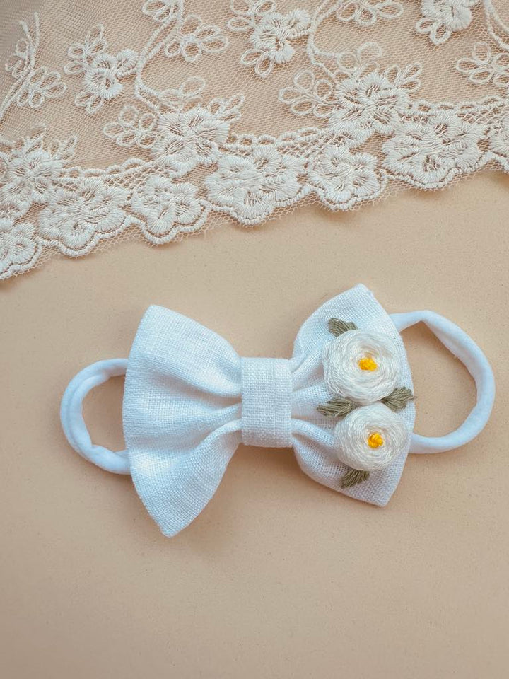 A white bow with flowers on it is placed on a beige background, with a lace doily visible in the top left corner.