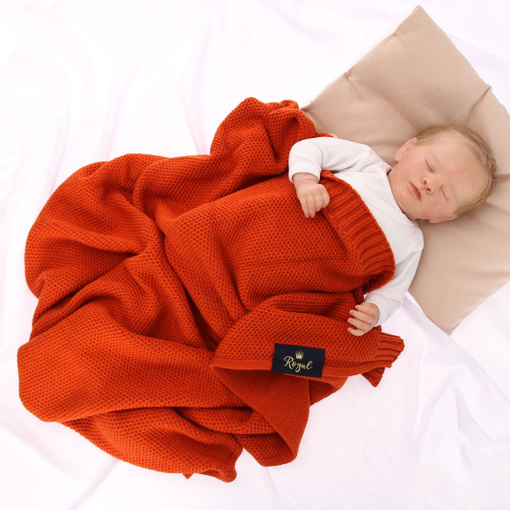 A newborn baby is peacefully sleeping, swaddled in a red blanket and resting on a beige pillow.