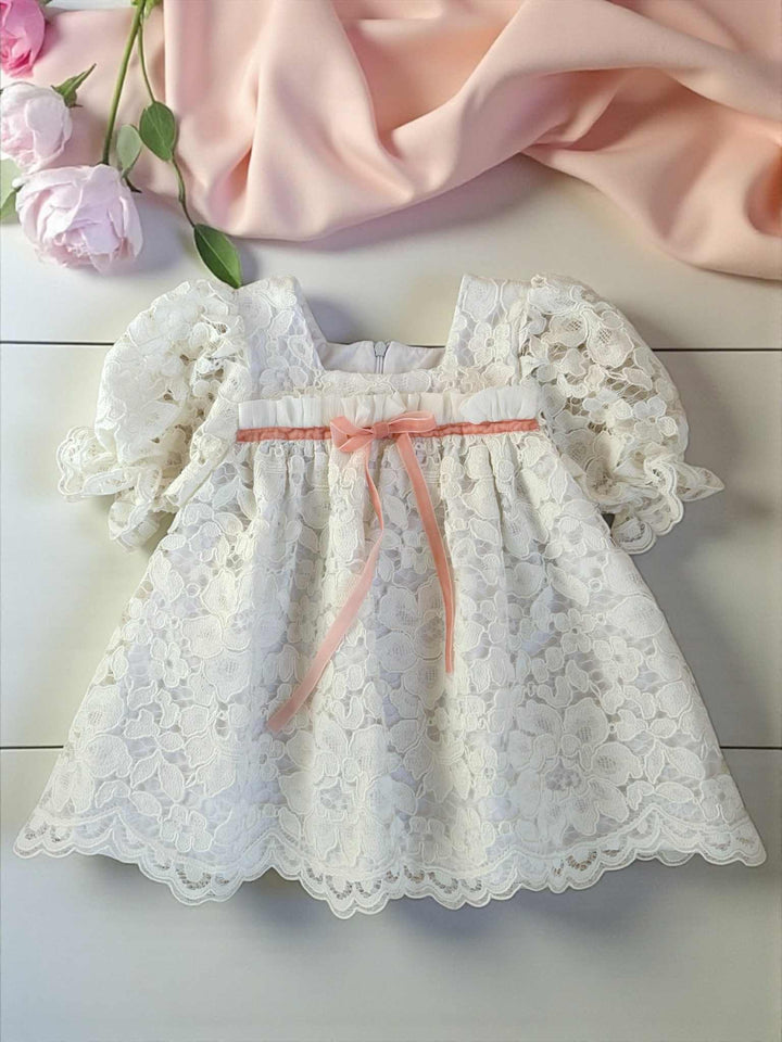 A white lace dress with a pink ribbon is displayed on a white surface, accompanied by pink roses and a pink fabric.