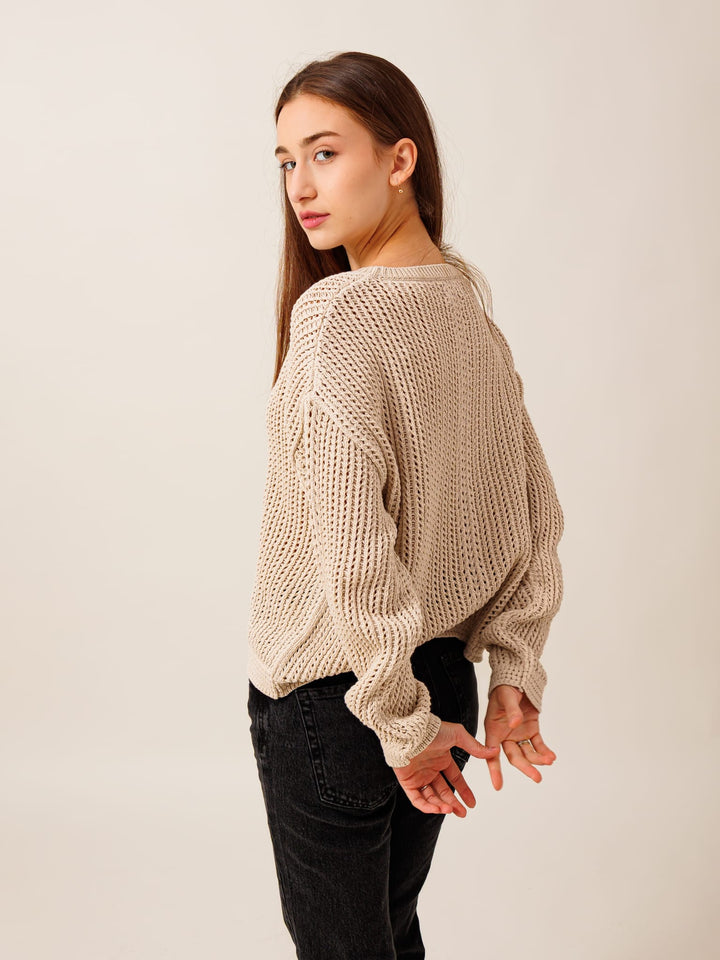 A young woman with long brown hair stands with her hands behind her back, wearing a beige sweater and black pants.