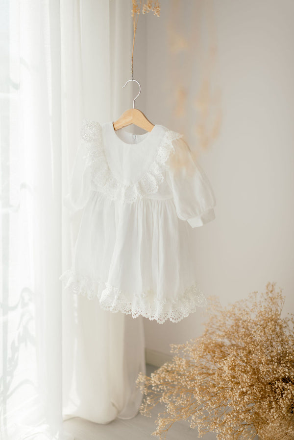 A white lace dress with ruffles is hanging on a wooden hanger in front of a window, with a bouquet of dried flowers on the floor next to it.