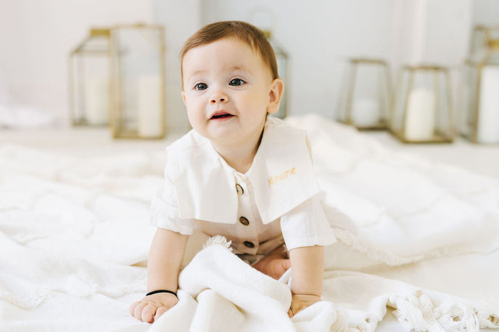 A smiling baby boy in a white outfit with a name tag is sitting on a white blanket, surrounded by candles in the background.