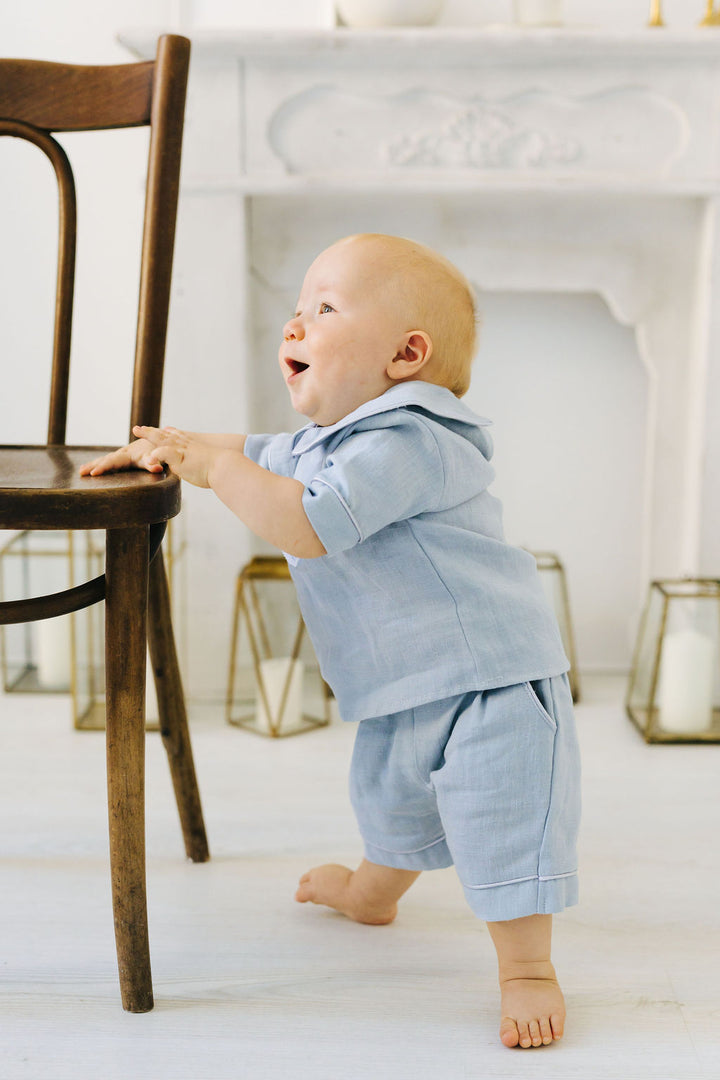 A baby in a light blue outfit is standing on a wooden chair, looking up with a joyful expression.