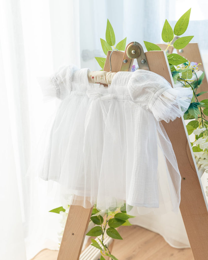 A white, ruffled dress is displayed on a wooden easel, surrounded by green leaves and flowers, creating a serene and elegant setting.