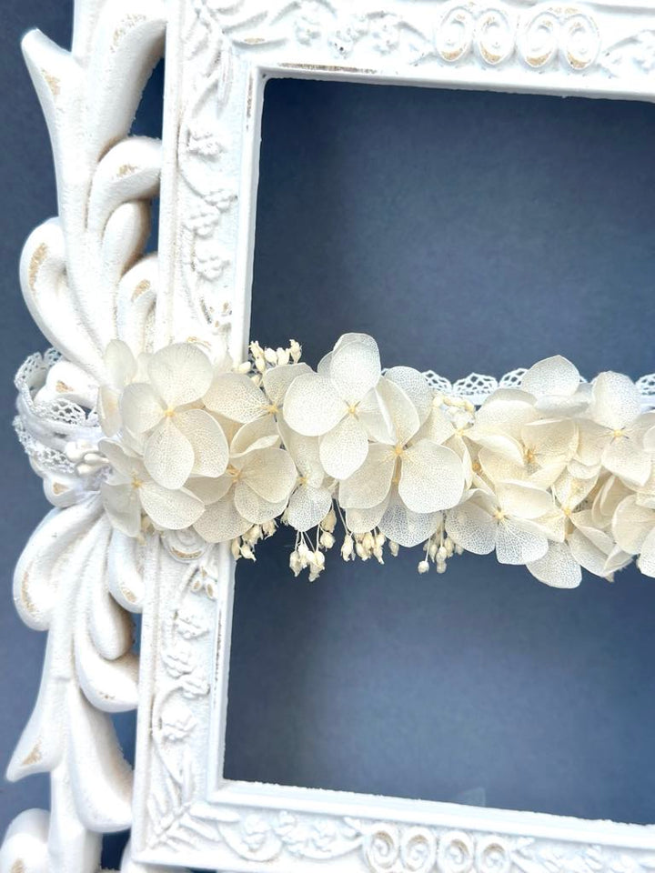 A white ornate frame with a floral arrangement of white flowers and lace trim.