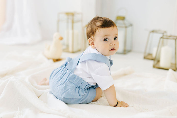 A baby boy in a white shirt and blue overalls is sitting on a white blanket, looking directly at the camera.