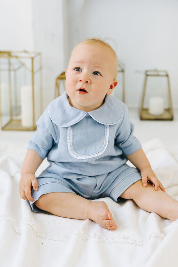 A baby boy wearing a light blue outfit sits on a white blanket, looking up with a curious expression.