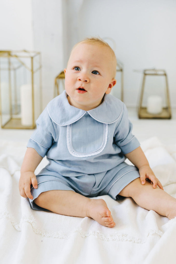 A baby boy wearing a light blue outfit sits on a white blanket, looking up with a curious expression.