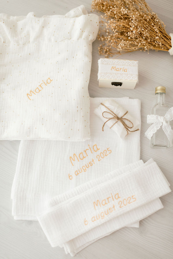 a variety of items related to a baby shower, including a white cloth with the name "Marie" embroidered on it, a small box, a bottle, and some dried flowers.