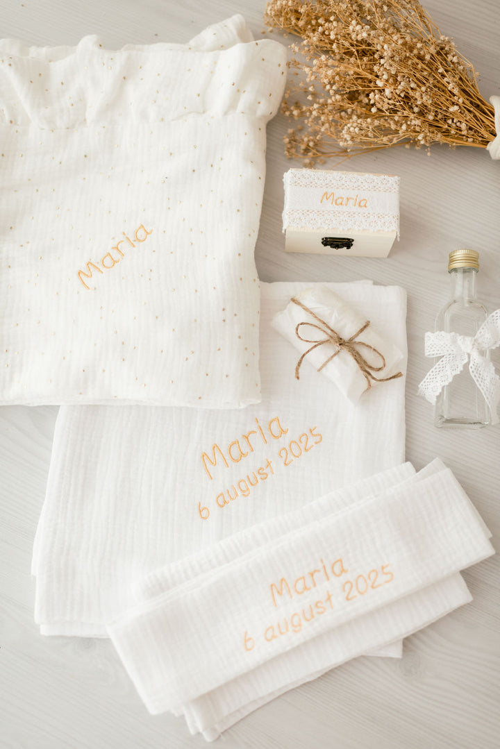 a variety of items related to a baby shower, including a white cloth with the name "Marie" embroidered on it, a small box, a bottle, and some dried flowers.