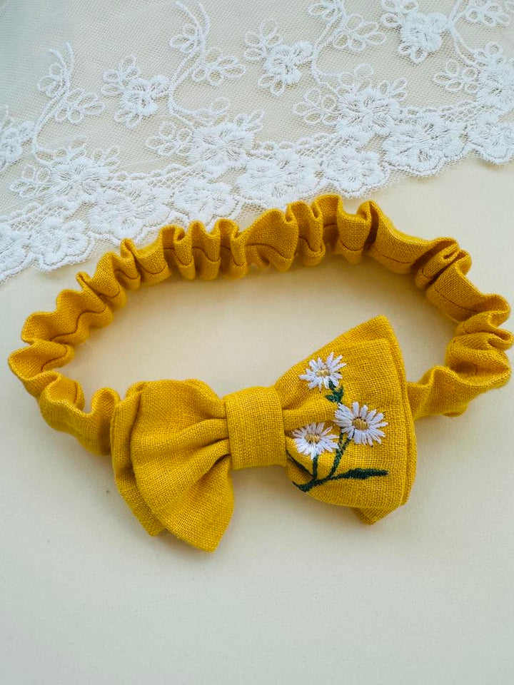 A yellow bow tie with white daisies embroidered on it, placed on a white lace background.