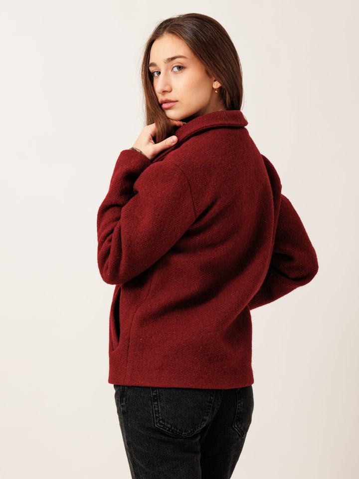 A young woman with long brown hair wearing a burgundy jacket stands with her hands on her hips, looking directly at the camera.