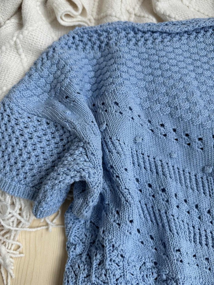 a close-up view of a blue knitted sweater with a textured, perforated pattern. The sweater appears to be made of a soft, light blue material and is resting on a wooden surface.