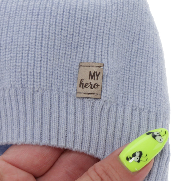 A close-up of a gray knitted hat with a small label attached to it, and a person's hand with neon green nail polish featuring a cartoon character.