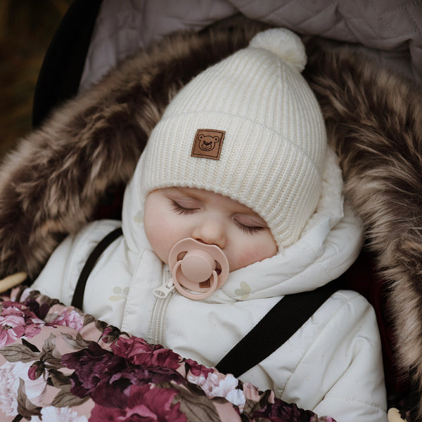 A baby sleeping peacefully in a white snowsuit and a white knit hat with a brown bear logo.