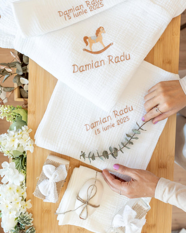 a person's hands arranging white towels with text and a horse embroidered on them, along with some decorative elements such as a plant and a ribbon.