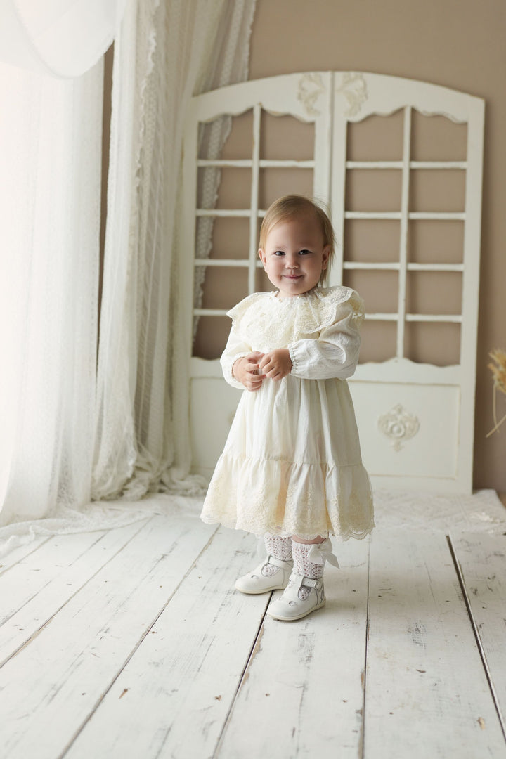 A young girl in a white lace dress stands in a white room with a wooden floor, smiling at the camera.