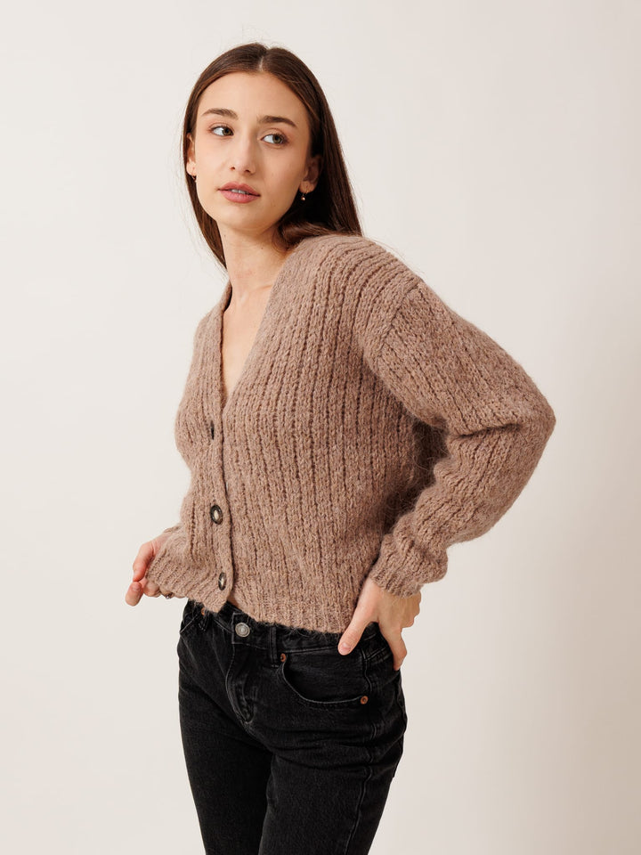 A young woman wearing a brown cardigan and black pants stands with her hands in her pockets, looking to the side.