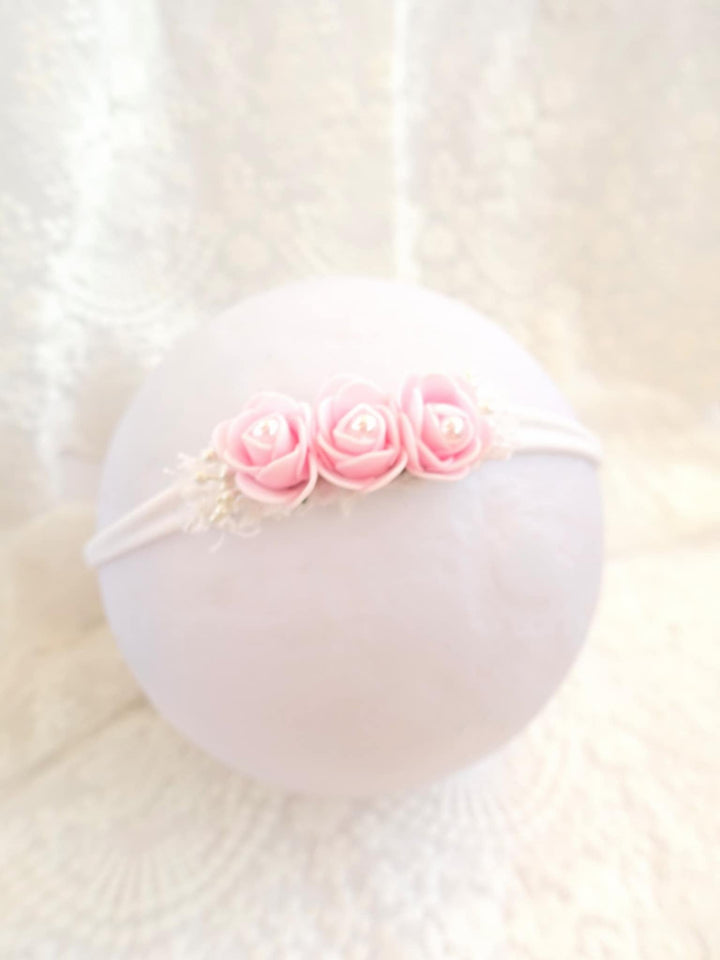 A white headband adorned with three pink roses is displayed against a white lace background.