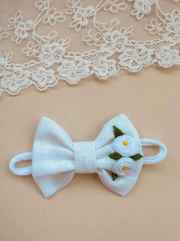 A white bow tie with two white flowers on it, placed on a beige background with a lace doily.