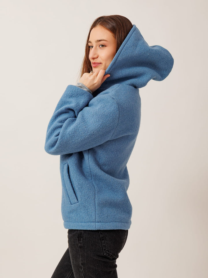 A woman wearing a blue hooded sweatshirt stands against a plain background, with her chin resting on her hand.
