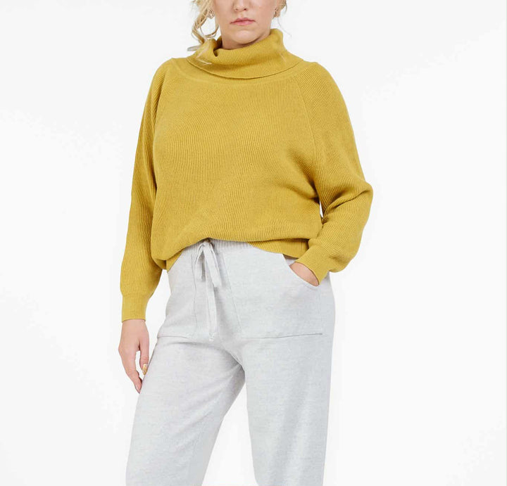 A woman wearing a yellow sweater and gray sweatpants stands against a white background.