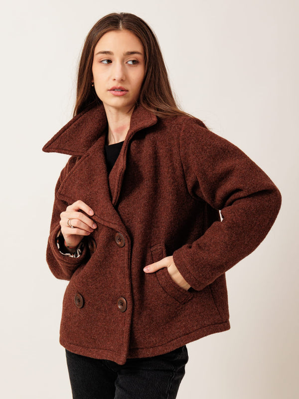A woman wearing a brown tweed coat stands against a plain background, looking off to the side with a serious expression.