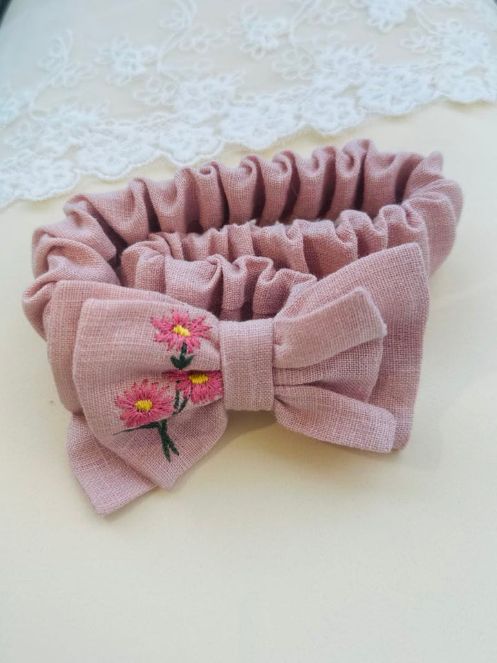 A pink bow tie with a floral embroidered design is displayed on a white surface, with a lace doily visible in the background.