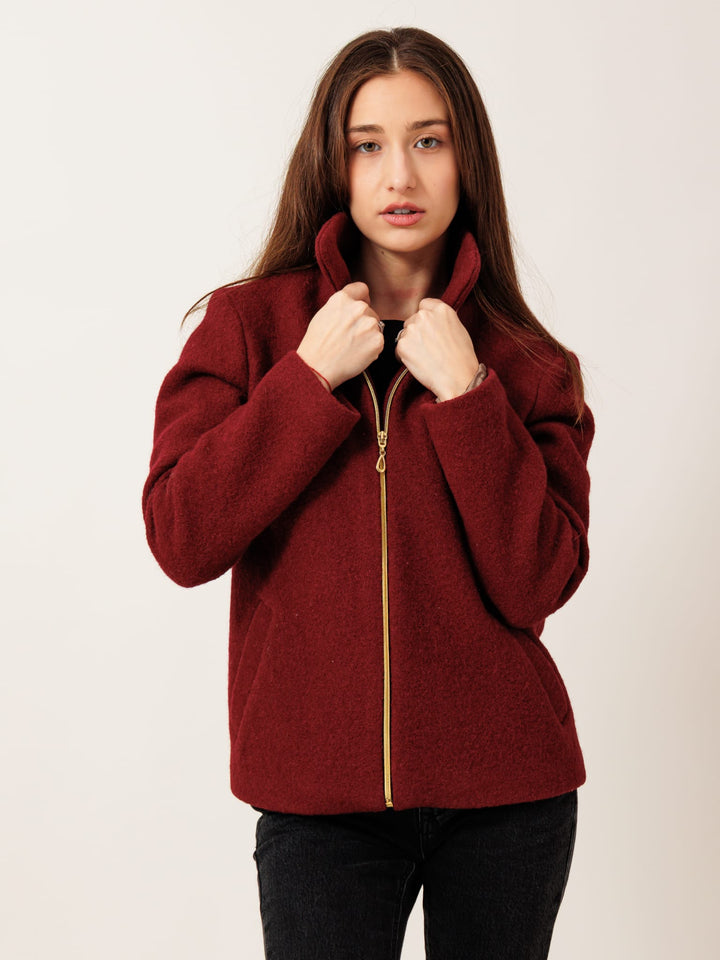 A young woman with long brown hair wearing a red jacket with a gold zipper stands against a plain white background.