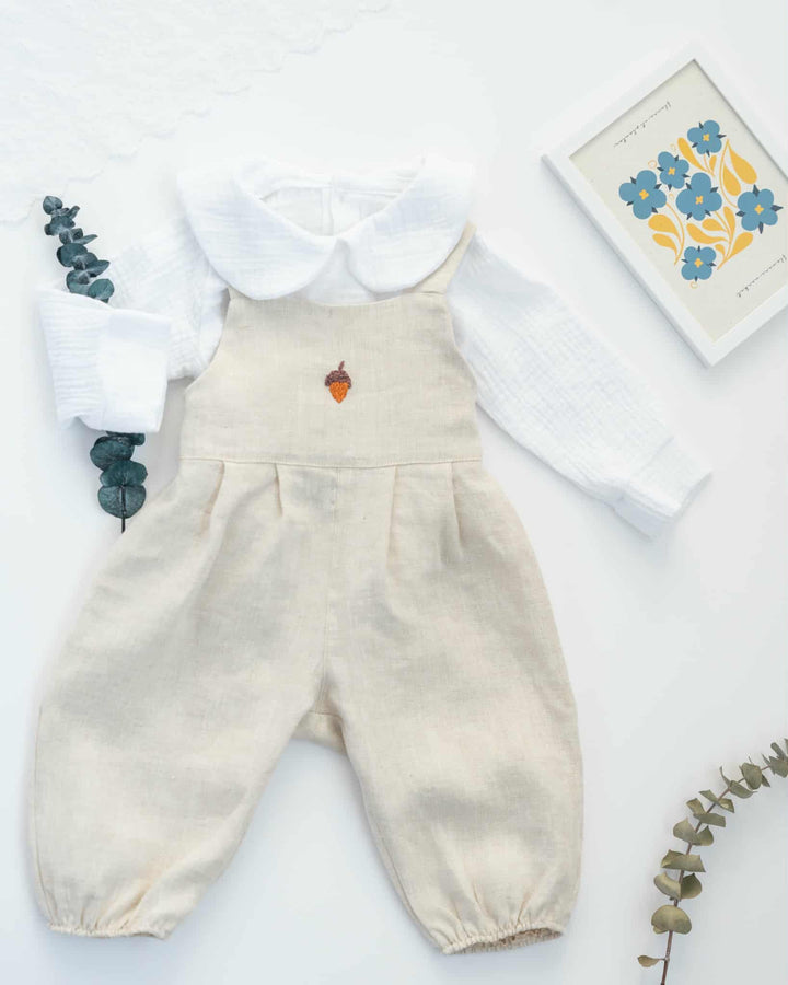 A beige outfit consisting of a white shirt and a white blouse with a white collar, featuring a small embroidered design on the front, is displayed on a white background alongside a framed floral print.