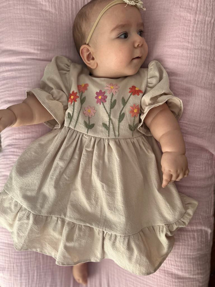 A baby girl wearing a beige dress with colorful floral embroidery is lying on a pink blanket.