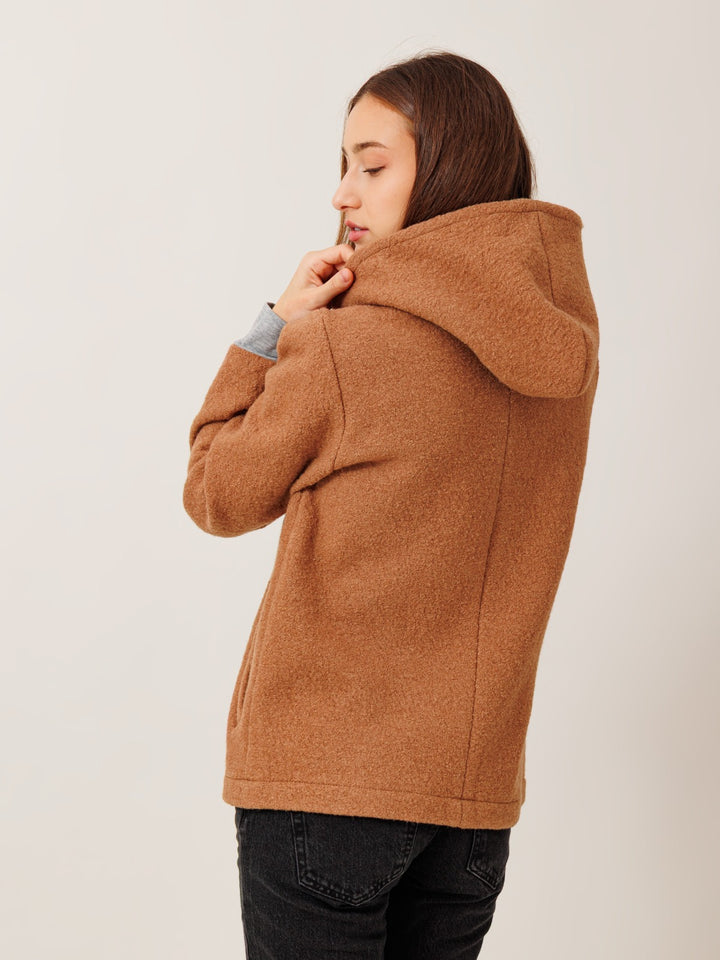 A woman wearing a brown hooded jacket stands with her back to the camera, her gaze directed downwards.