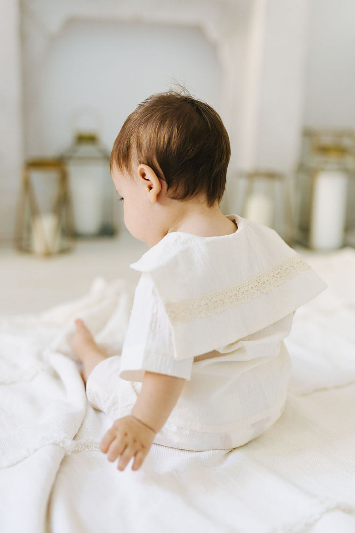 A baby in a white outfit is sitting on a white blanket, facing away from the camera.