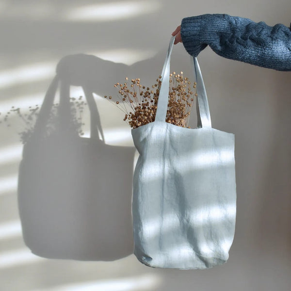A hand holding a white tote bag with dried flowers inside, against a plain white wall.