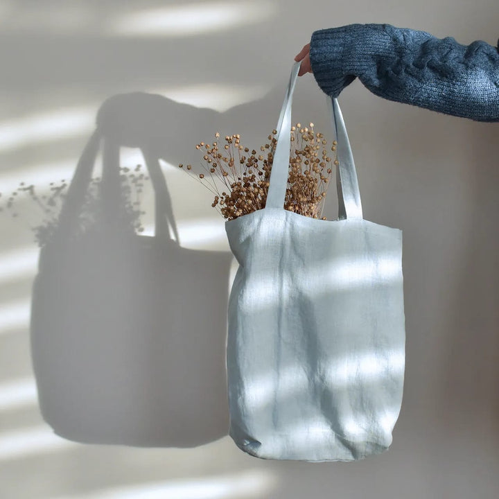 A hand holding a white tote bag with dried flowers inside, against a plain white wall.