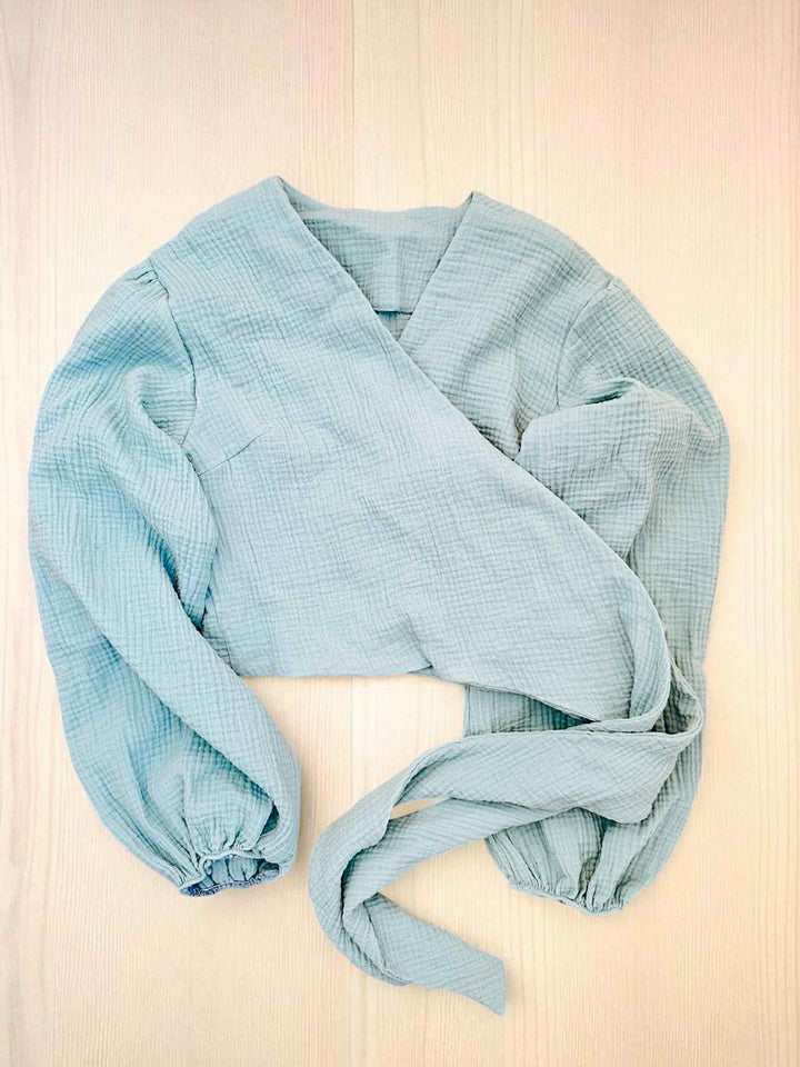 A light blue, long-sleeved blouse with a wrap-style neckline is displayed on a light wooden surface.