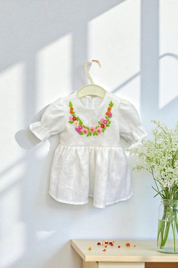 A white dress with colorful floral embroidery is hanging on a white hanger against a white wall, accompanied by a vase of white flowers on a wooden table.